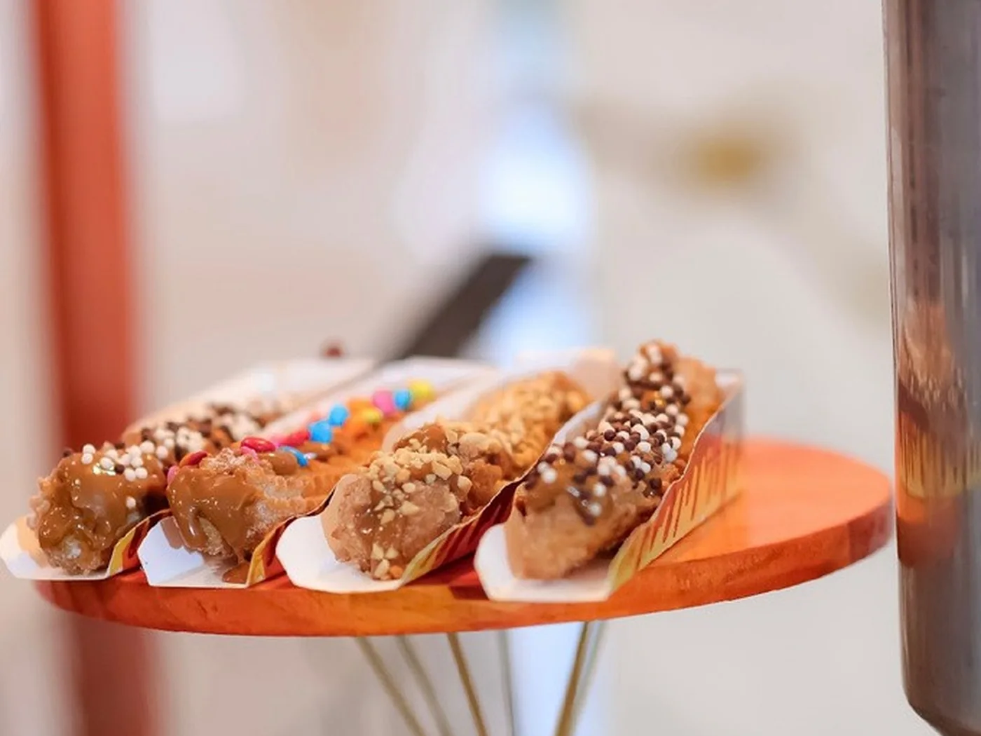 Churros gourmet cobertos com chocolate e complementos dispostos em suporte de madeira